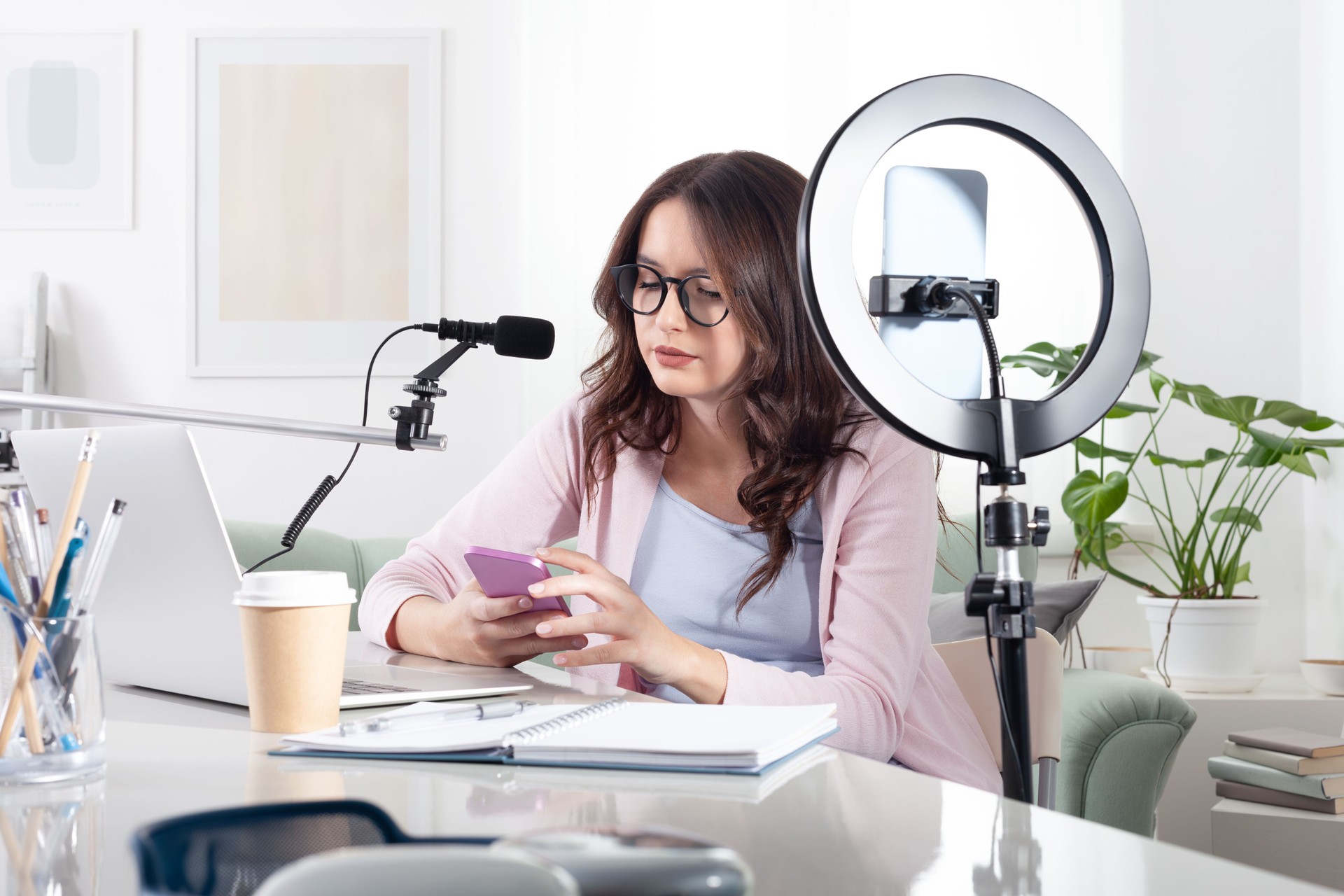 Smiling female content creator and influencer at home with smartphone and ring light, scrolling on her phone. Live streaming or recording a tutorial or social media content for her online followers